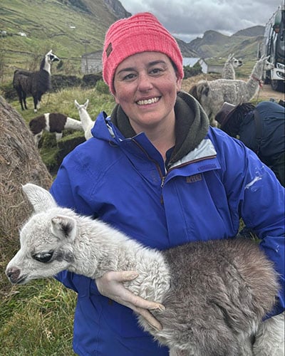 A smiling veterinarian in a pink beanie and blue jacket holds a baby llama in a grassy field, surrounded by several llamas and rolling hills in the background.
